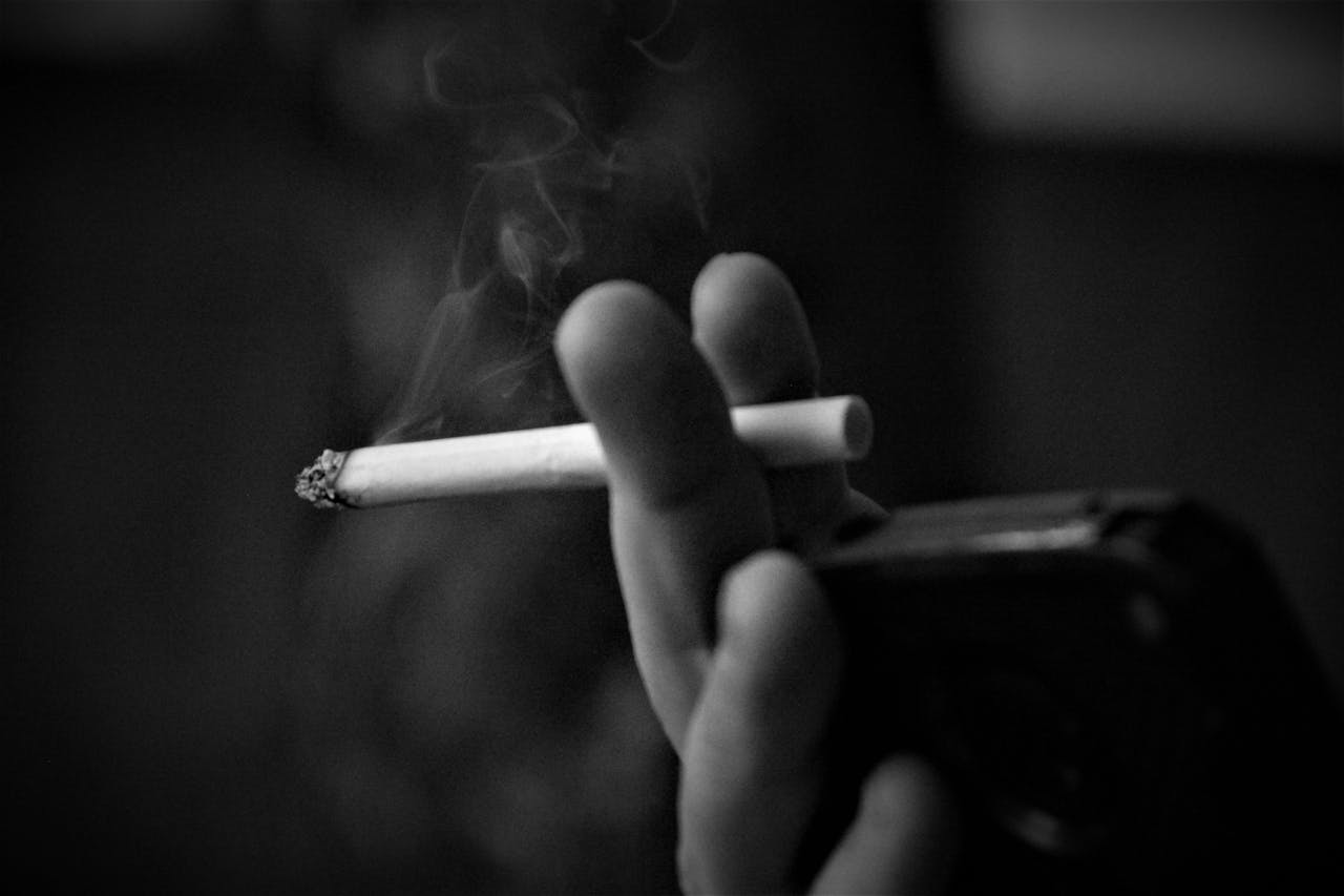 Black and white close-up of a hand holding a lit cigarette with smoke rising against a dark background.