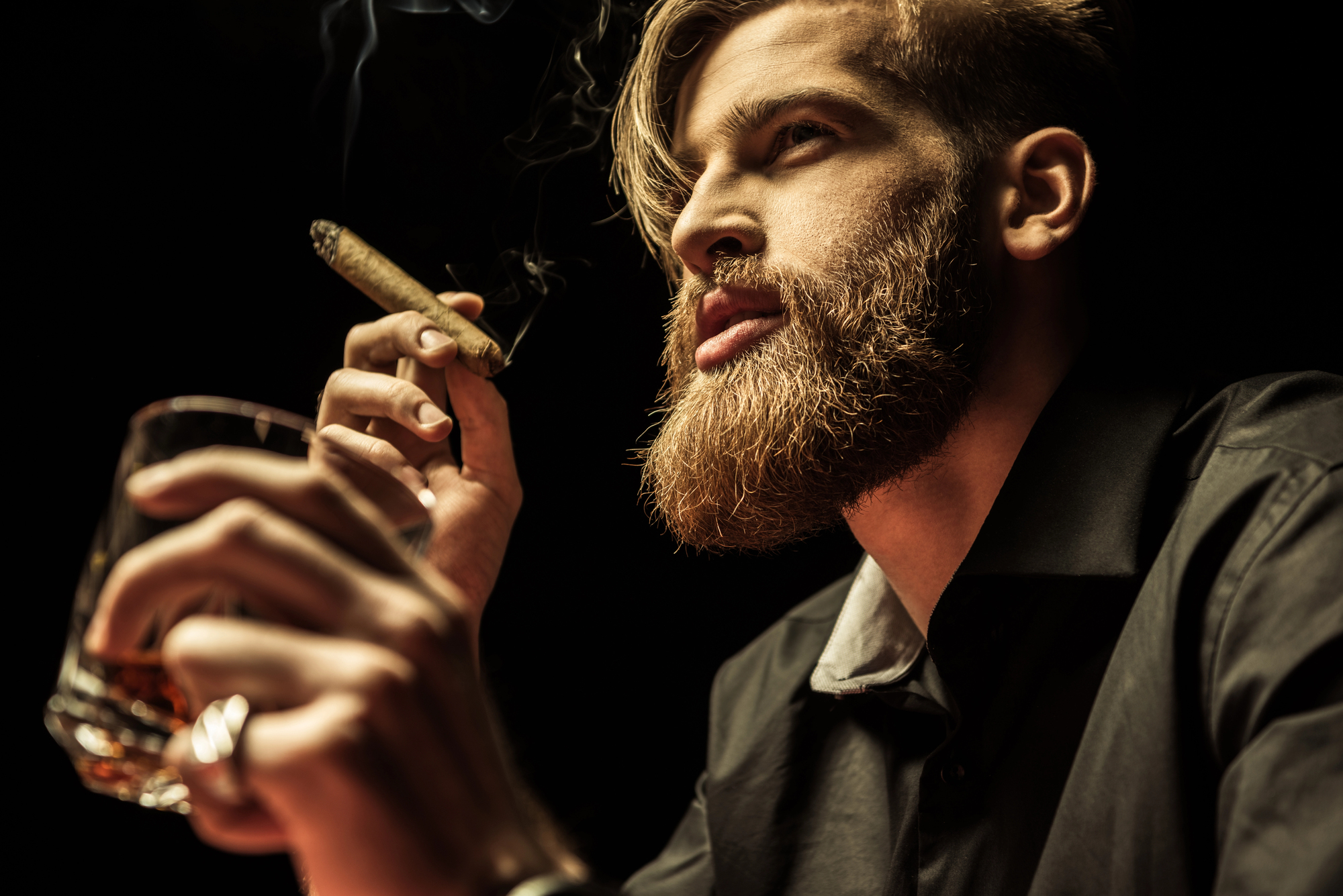 Bearded man holding a lit cigar and glass of whiskey, gazing upward against a dark background.