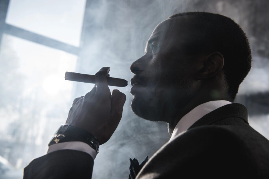 Man in black suit smoking a cigar surrounded by smoke, shot from below in dramatic low-key lighting.